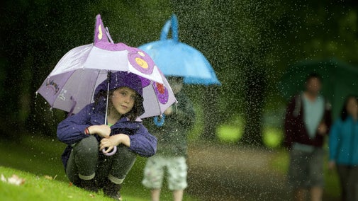 Two children crouch under umbrellas from the rain in a green wooded area. The rain is slightly misty and out of focus in front of them